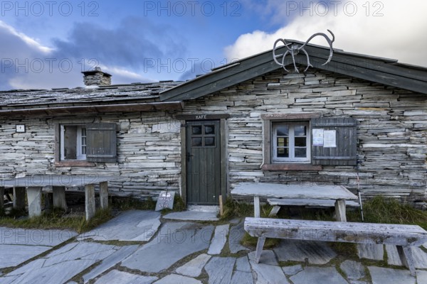 A traditional stone house with a café sign, wooden benches in front, surrounded by a mountainous