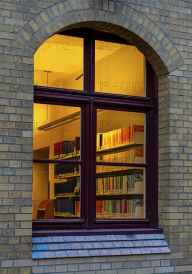 A view through the window of a library, shelves full of books illuminated by a warm, yellow light,
