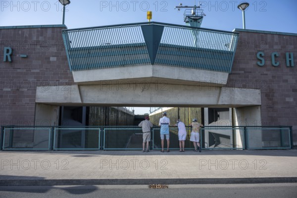 Four people stand in front of a modern lock structure made of concrete and blue railings, Minden