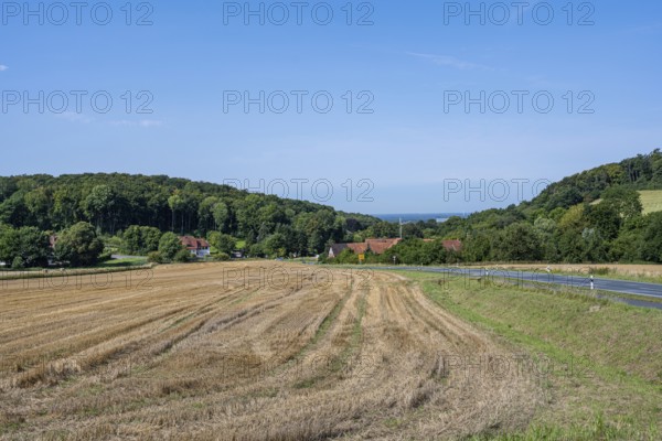 View of a harvested field with neighbouring village and wooded hills under a clear sky, Halle,