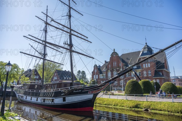 A historic sailing ship on a sunny day with a striking brick building in the background, The