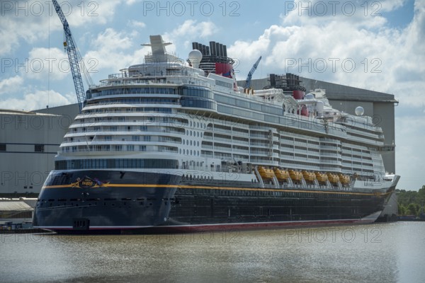 A large cruise ship lies calmly in the water next to a large shipyard on a partly cloudy day,