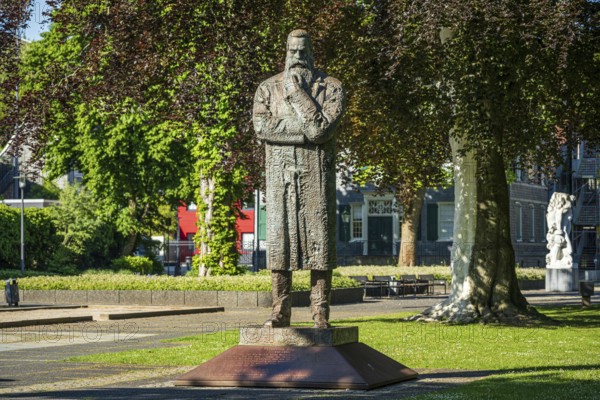 Stone statue of a man with crossed arms in an urban park with surrounding buildings, statue of