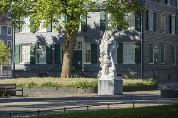 Stone figure in front of a historic building with green shutters, surrounded by blossoming trees,