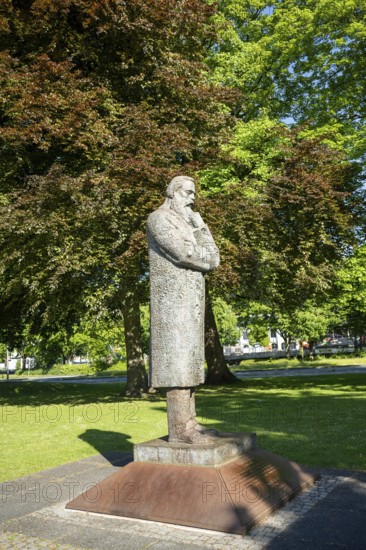 Side view of a stone statue of a pensive man in a green park with trees, statue of Friedrich Engels