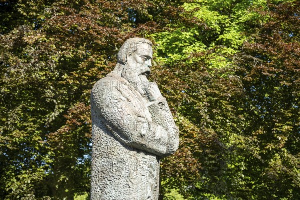 Stone statue of a pensive man surrounded by lush green trees in a park, statue of Friedrich Engels