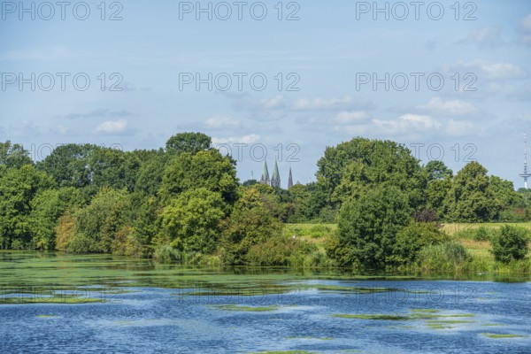 A river with green foliage, church towers in the background and blue sky, Weser cycle path, Weser,