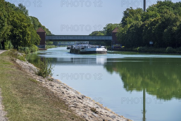 Cargo ship under a bridge, flanked by trees. Tranquil river landscape with greenery, Dortmund-Ems