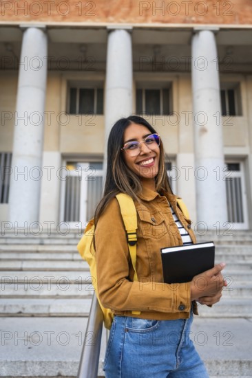 Young woman smiling, wearing glasses and a backpack, standing with a notebook in front of a
