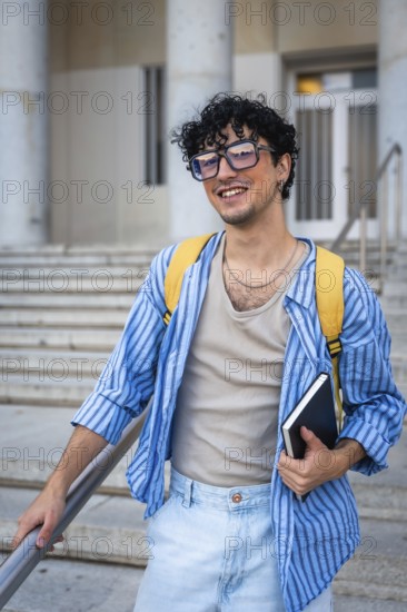 Young man with a backpack and a notebook, smiling while walking up the modern building stairs,