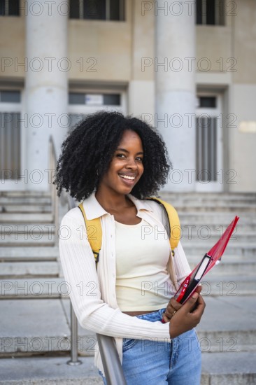 African american woman smiling while holding a notebook, wearing a backpack and standing in front