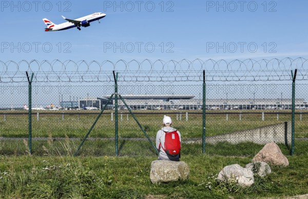 A man sits on a stone and watches the take-off of a British Airways plane through a barbed wire