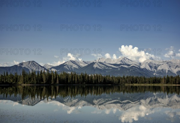 Tranquil lake mirroring mountains of the Canadian Rocky Mountains and sky, with pine forest and