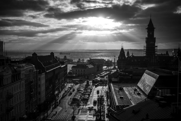 Black and white cityscape with dramatic clouds and sunlight over a harbor and buildings and looking