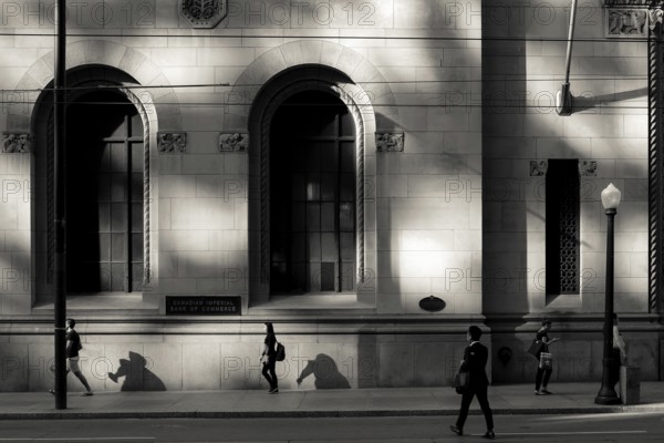 Black and white urban scene with people walking past a sunlit building casting strong shadows in
