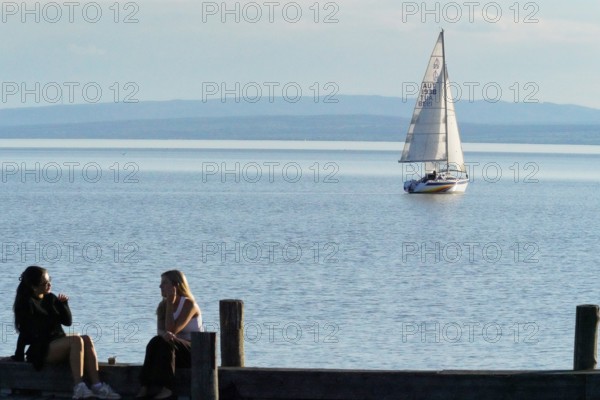 Lake Neusiedl, Burgenland, Austria, Europe, Two woman sit on the jetty while a sailing boat passes
