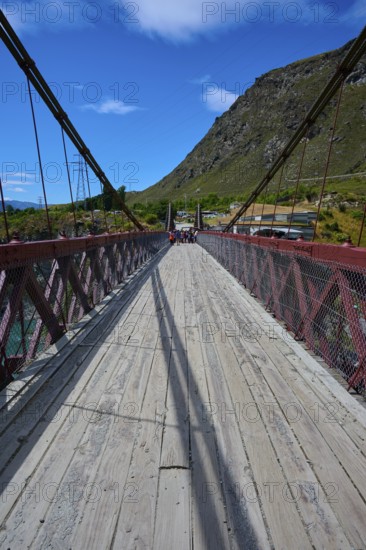 Wooden footbridge with red railing, shadows and mountain background, Kawarau Gorge Suspension
