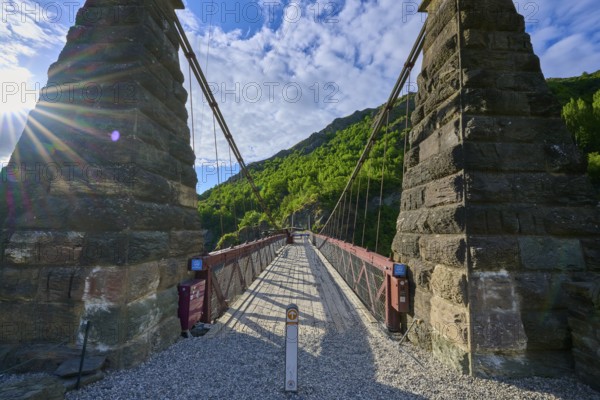 Old bridge with massive stone pillars surrounded by lush greenery under a bright blue sky, Kawarau