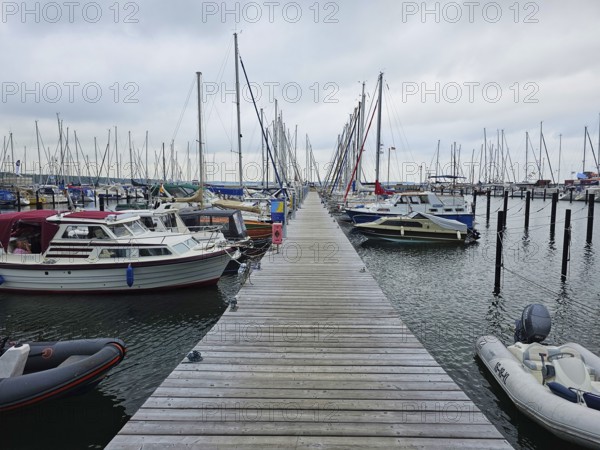 A wooden walkway leads through a harbour, lined with boats and masts under a cloudy sky, Laboe,