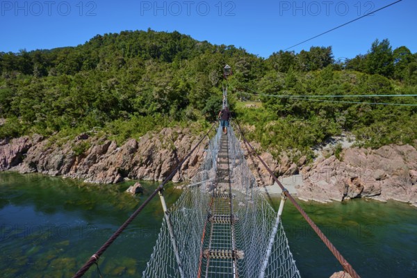Suspension bridge over turquoise river, surrounded by dense forest under blue sky, Buller Gorge