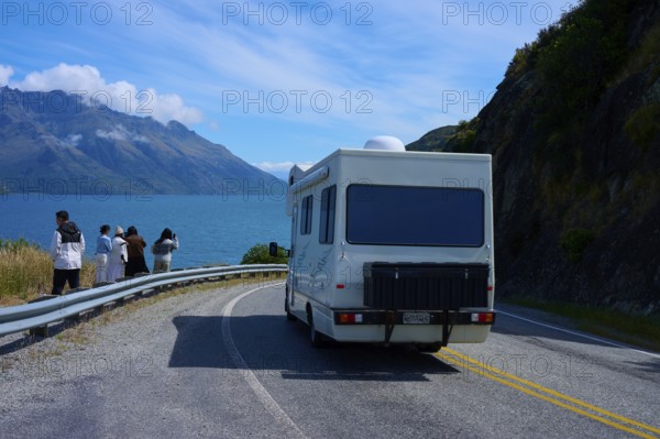 People enjoying travelling and a motorhome driving on a road along a lake surrounded by mountains,