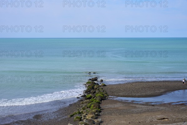 View of the calm sea with rocks leading into the water and a lone walker on the beach, Mana Bay,