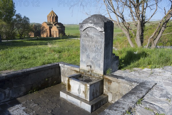 Stone fountain with engraving in front of a rural church in a green landscape, original spring,