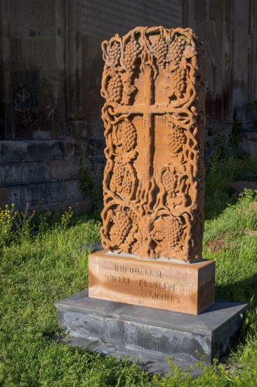 A detailed engraved red gravestone with a cross motif stands on a grassy area in the sunlight,