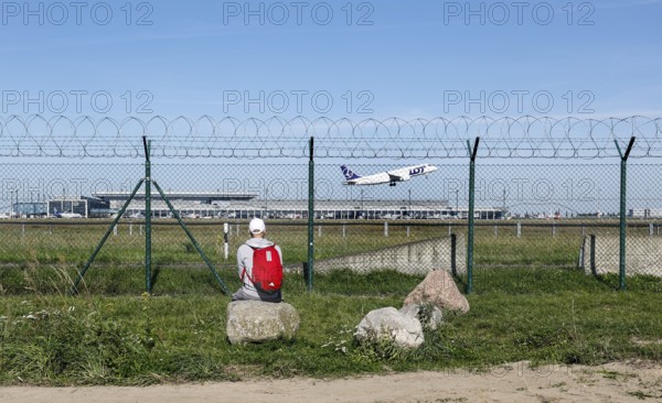 A man sits on a stone and watches an aircraft of the Polish airline LOT take off through a barbed
