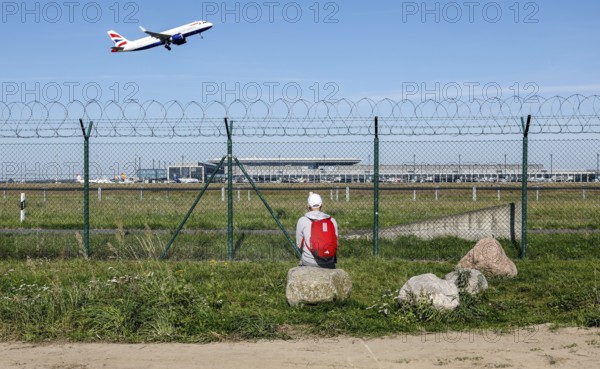 A man sits on a stone and watches the take-off of a British Airways plane through a barbed wire