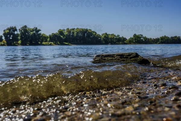 The sun shines on the calm waters of a river. Small waves gently lap against the shore, while the