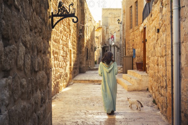 Woman in traditional abaya walking with cat through narrow stone alley in historic city of Mardin