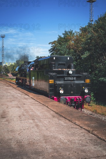 An old steam locomotive stands on the tracks next to an overgrown platform under a blue sky, Gera,