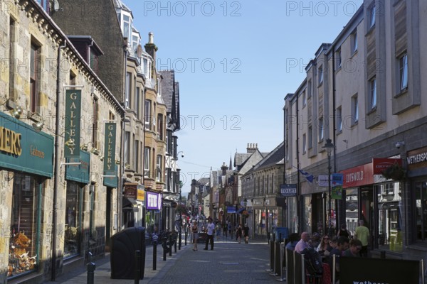 Busy shopping street with people and traditional buildings under a blue sky, Fort William,