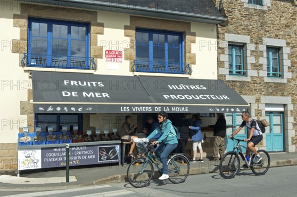 Cyclists on the Quai John Kennedy pass a shop selling seafood such as oysters and mussels, Cancale,
