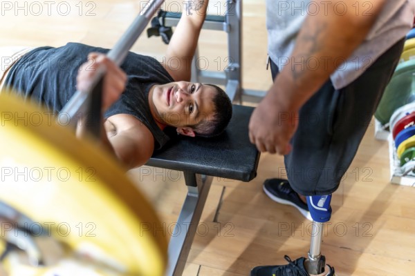 Disabled man performing a bench press with a barbell while a friend with a prosthetic leg stands