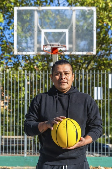 Indigenous man standing on an outdoor basketball court holding a bright yellow basketball, looking