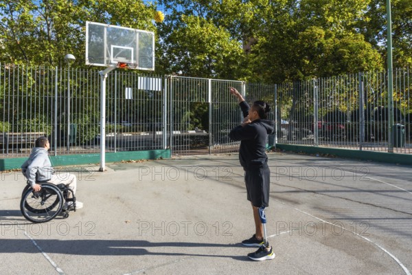 Two athletes playing basketball on an outdoor court. One sitting in a wheelchair and the other