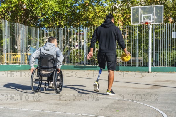 Man with prosthetic leg dribbling basketball on outdoor court while friend in a wheelchair watches,