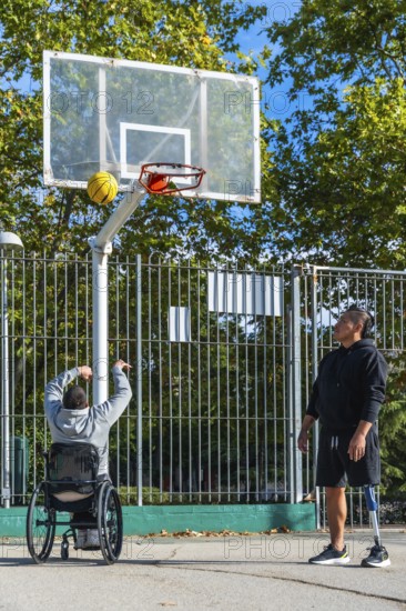 Two men play outdoor basketball, one shooting from a sports wheelchair while the other stands with