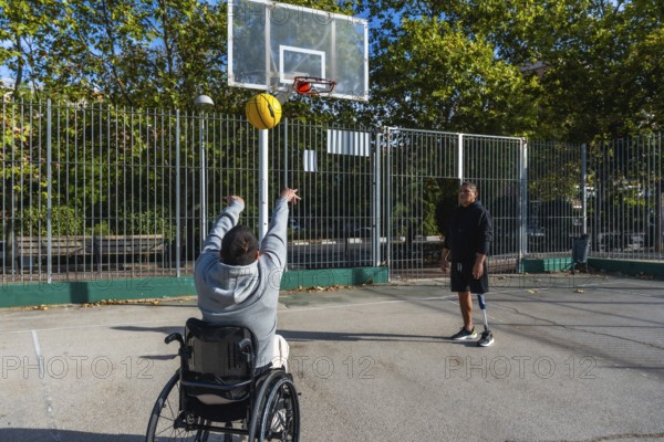 Two young men playing basketball outdoors, one in a wheelchair shooting the ball towards the hoop