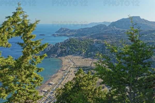 View of the Tsambika sandy beach, with mountains and blue sea, framed by green trees, Church of the
