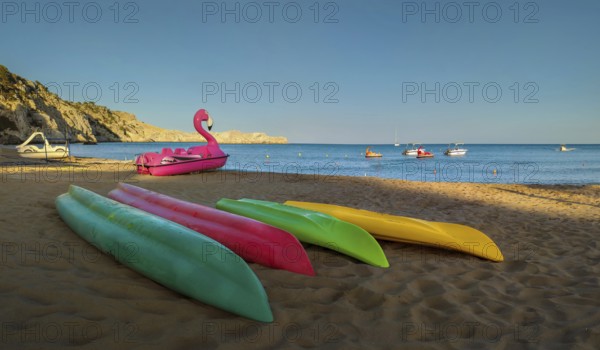 Colourful kayaks lying on Tsambika sandy beach, next to a pink flamingo pedal boat, boats floating