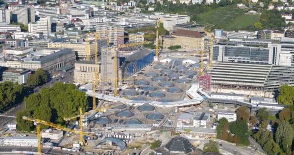 View of an urban construction site with cranes and modern glass structures, surrounded by green