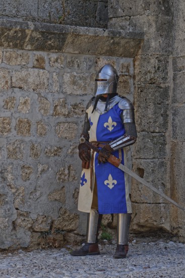 A knight in medieval armour stands with a sword in front of a stone wall, Medieval Town of Rhodes,