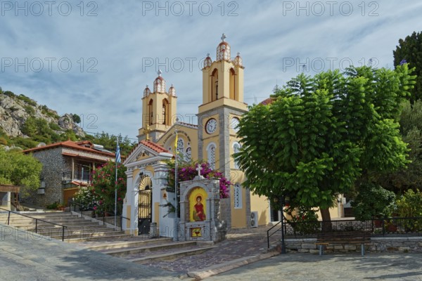 A yellow church with towers and bells surrounded by trees under a cloudy sky, Saint Pantaleon Greek