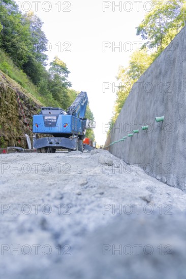 Blue excavator on a path next to a concrete wall with green pipes, Hermann Hessebahn construction