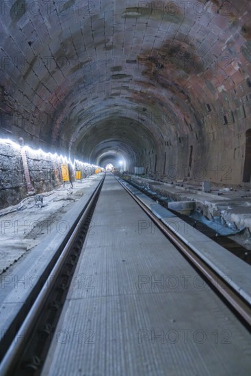 A long, illuminated tunnel with tracks and a deep perspective. The walls appear structured,