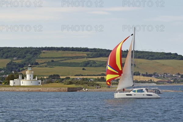 A sailing boat with a colourful sail sails near a lighthouse on the coast, Moray Firth, Inverness,