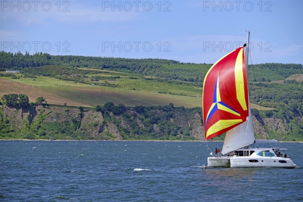 A sailing boat with a vividly coloured flag sails in front of a mountainous coastal landscape,